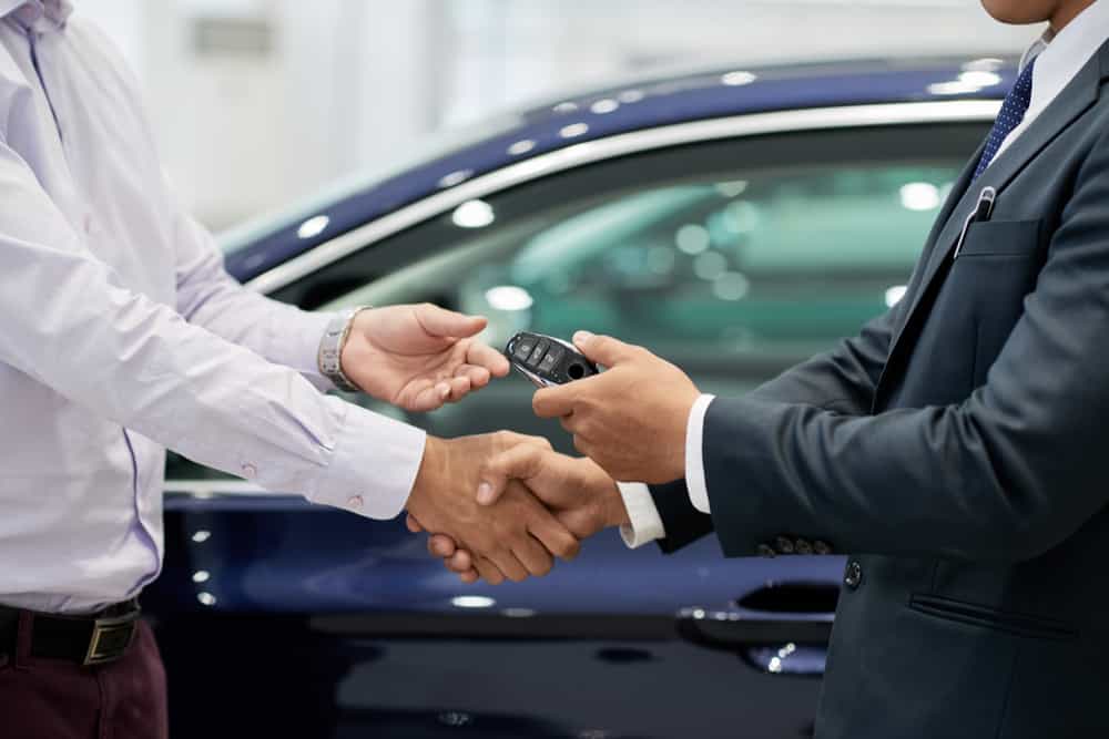 Comercial de concesionario estrechándole la mano a un hombre mientras le da las llaves de un coche
