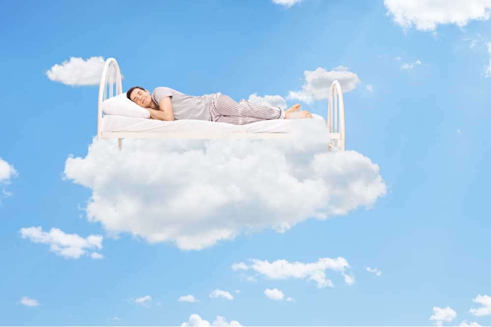 joven durmiendo feliz en una cama sobre una nube en el cielo azul
