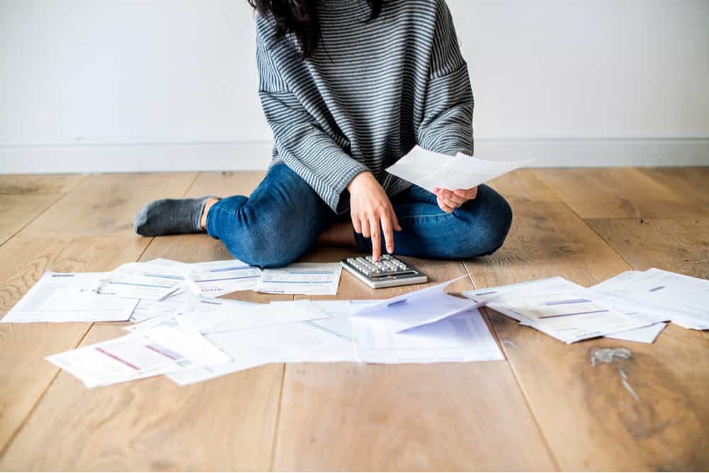 Chica sentada sobre el suelo rodeada de un montón de facturas en papel, usando una calculadora