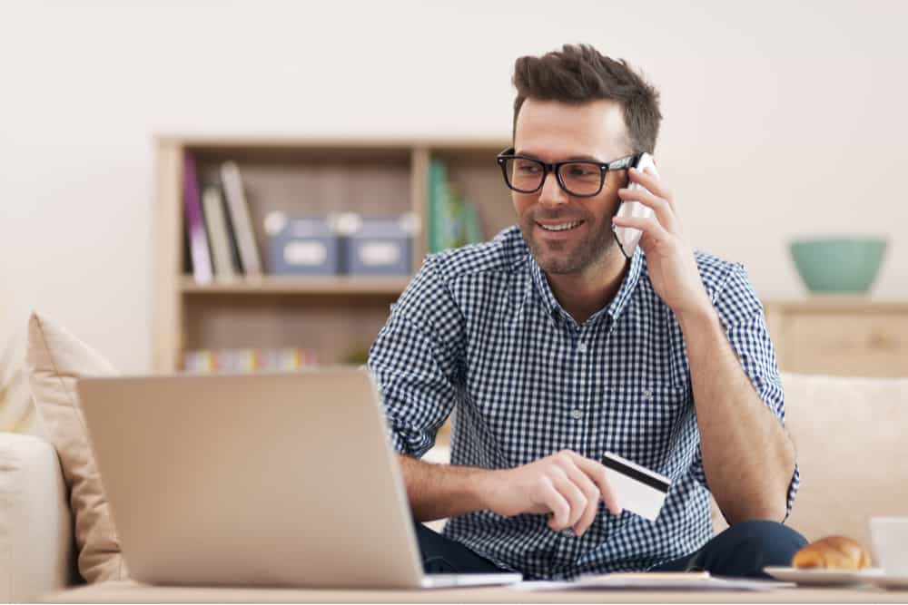 Hombre con gafas y camisa de cuadros sentado en el sofá hablando por el móvil con el portátil sobre la mesa y una tarjeta de crédito en la mano