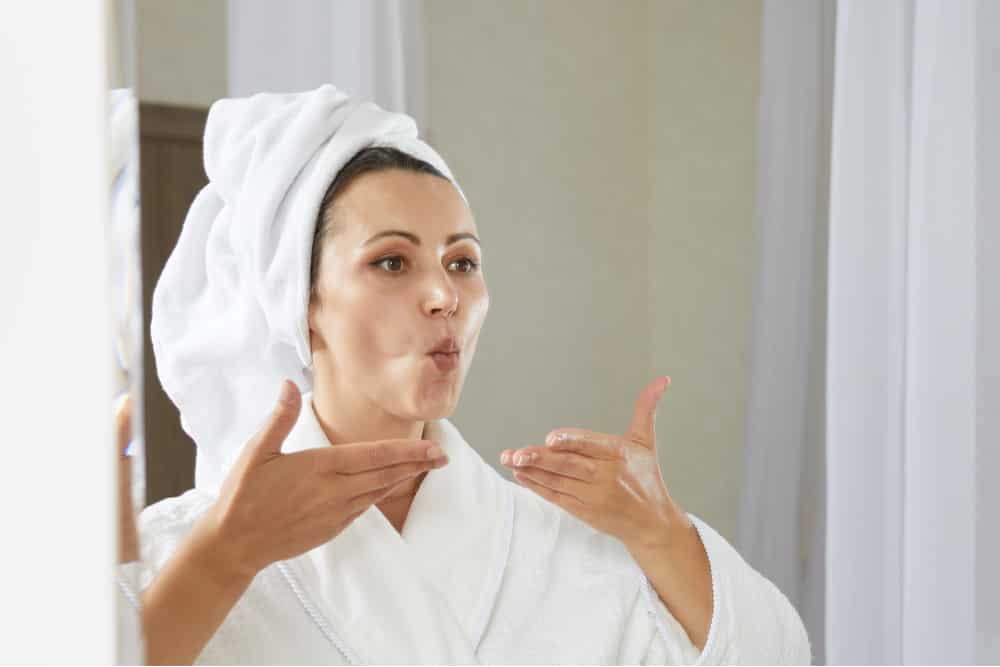Mujer con albornoz y turbante realizando yoga facial frente al espejo